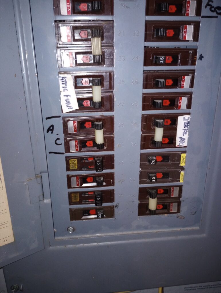 An open electrical panel showing multiple circuit breakers with amperage ratings (15A, 20A, 30A, 50A) and handwritten labels including 'AC' and 'Attic Fan'.