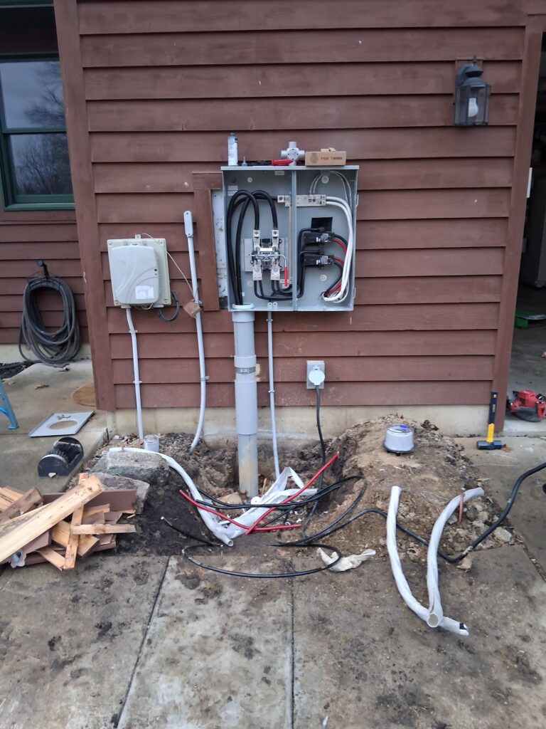 An open electrical meter base and service panel mounted on a brown wood-sided house, with exposed wiring and conduit running into a dug-out area with cables during an installation or upgrade.