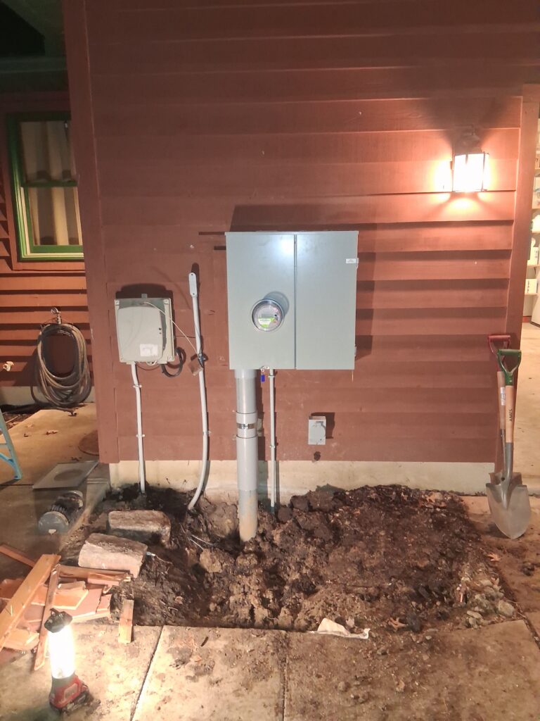 A newly installed electrical meter and service panel mounted on a reddish-brown wood-sided building at night, with a conduit running down into freshly dug soil, surrounded by construction tools including shovels, a lantern, and a coiled hose.