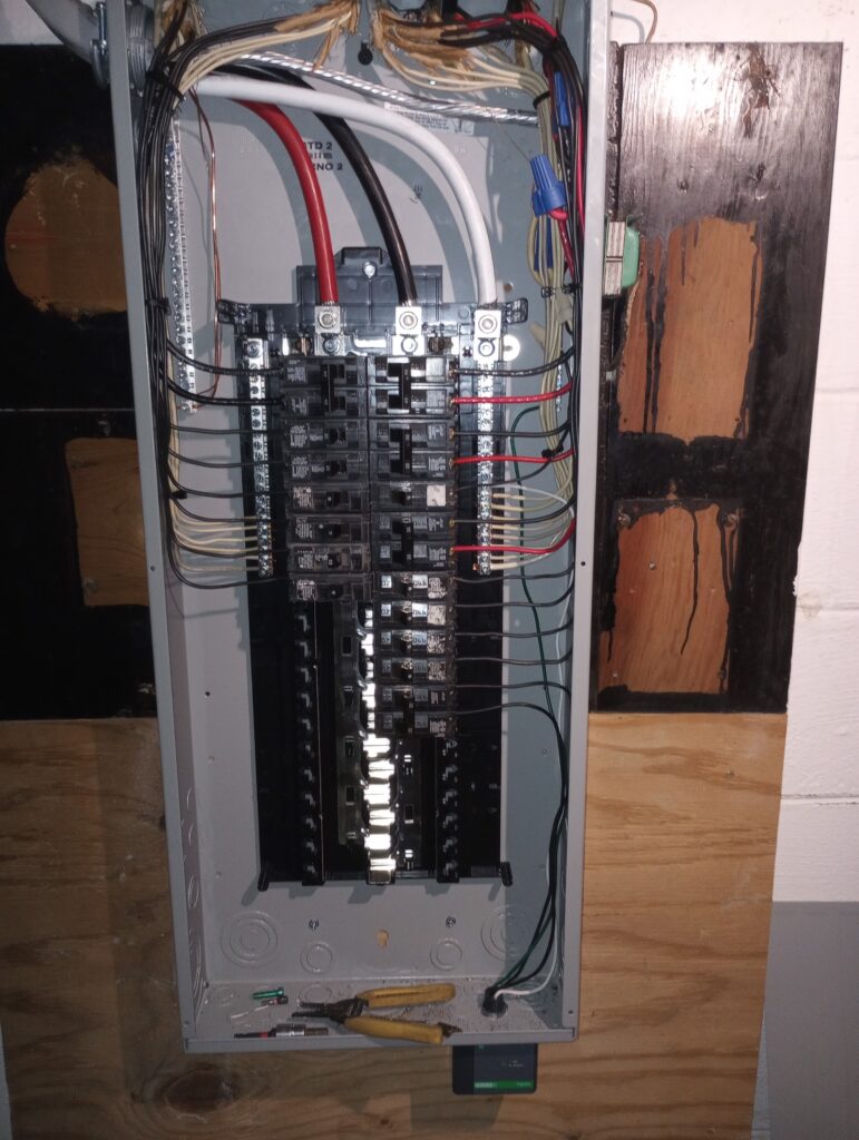 An open residential electrical breaker panel showing circuit breakers, colored wiring, and service cables, mounted on plywood with fire-damaged wall nearby and tools resting inside the panel box.